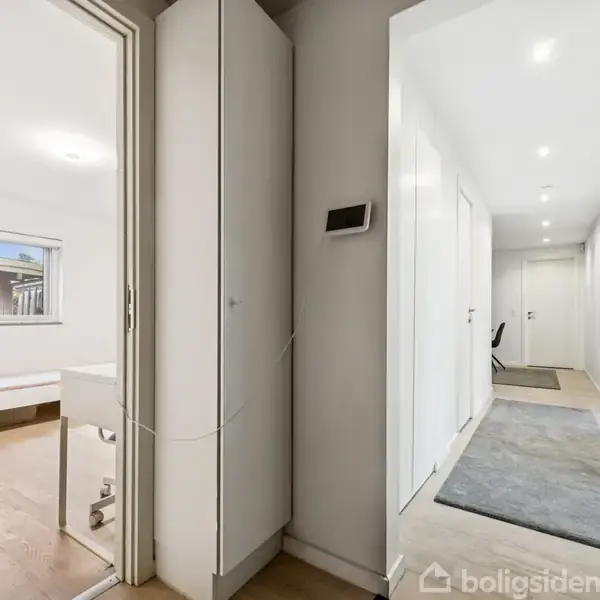 En lys gang med trægulv og hvide vægge fører til flere døre. Til venstre ses et værelse med en seng og udsigt til en gårdhave gennem et vindue.