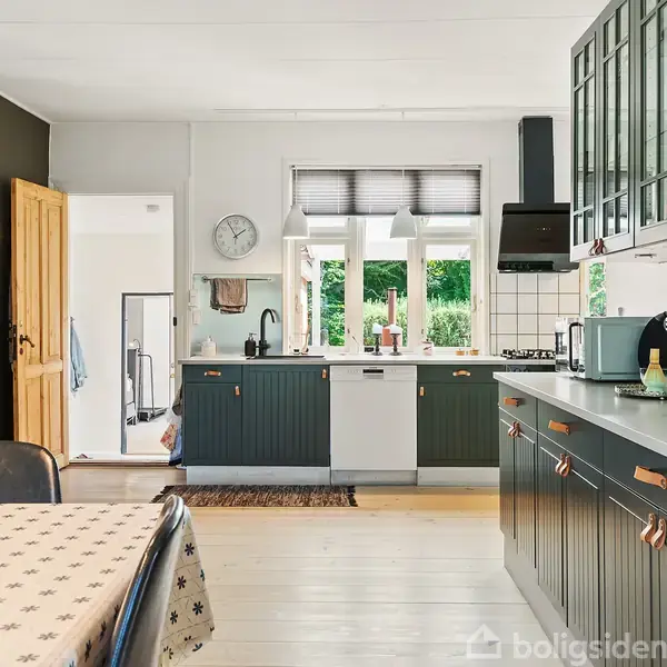 Køkken med grønne skabe udfører opbevaring. Bordet er dækket med en mønstret dug, og en lampe hænger over det. Vinduer giver udsigt til grøn natur udenfor.
