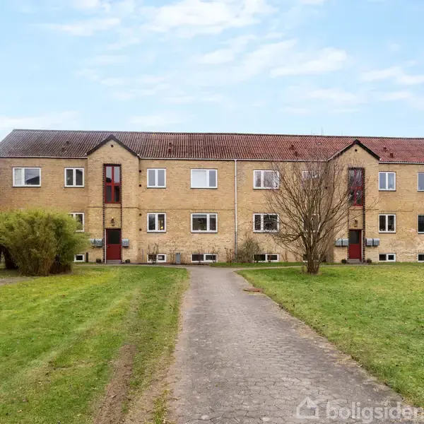 To-etagers lejlighedskompleks i gule mursten står stille, omgivet af en grøn græsplæne med træer. En lang, brolagt sti fører op til bygningen. Ingen tekst er synlig.