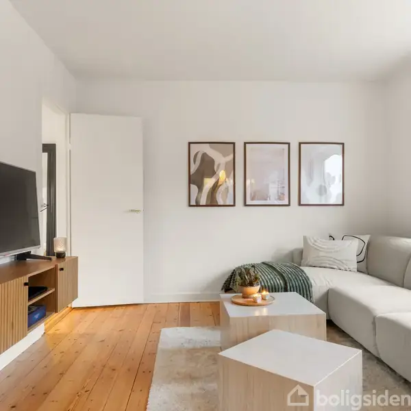 Stue med hvid sofa i hjørne, stor tv-skærm på et træbord og tre kunstværker på hvid væg. Trægulv og lyst indfald fra et vindue til højre.