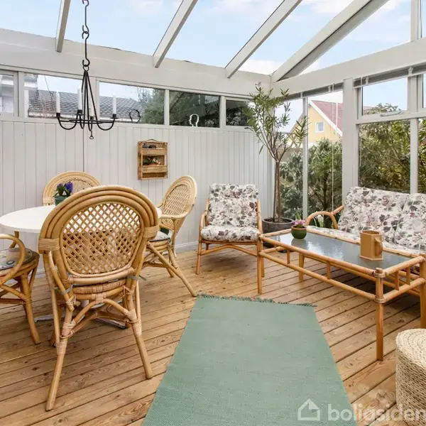 Fletmøbler placeret i udestue, herunder bord og stole samt sofaer med blomstrede hynder. Træterrassegulv og glasloft omslutter rummet med udsigt til have og husfacade.