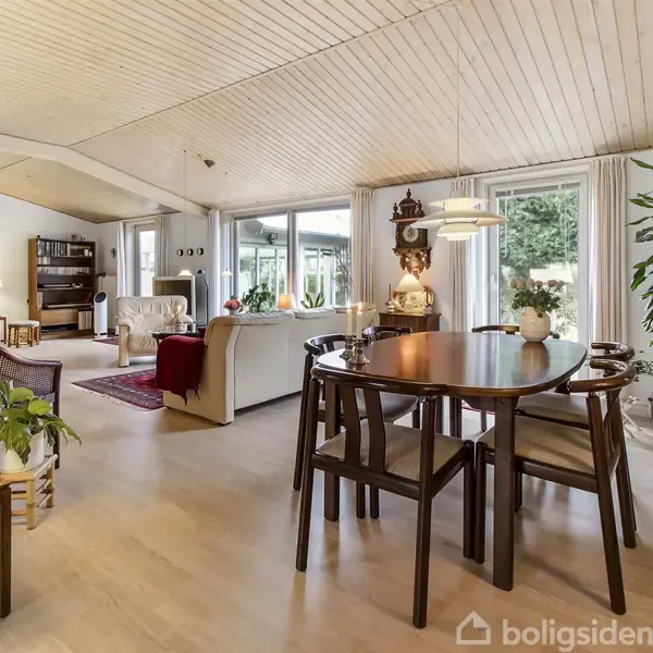 Spisebord med stole står midt i en stue. Stuen er rummelig med lyse vægge og træloft, indeholder sofaer, lamper, planter og store vinduer, der giver udsigt til haven udenfor.