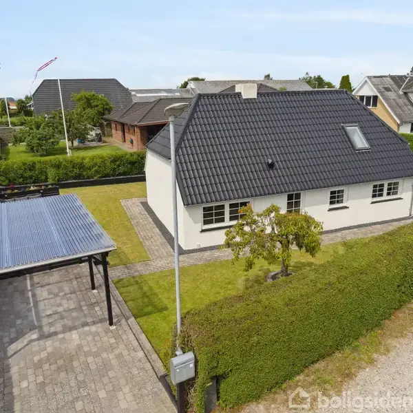 Et hvidt hus med sort tegltag står i en grøn have med en enkelt lille træ. En carport med transparent tag er på venstre side, omgivet af hække.
