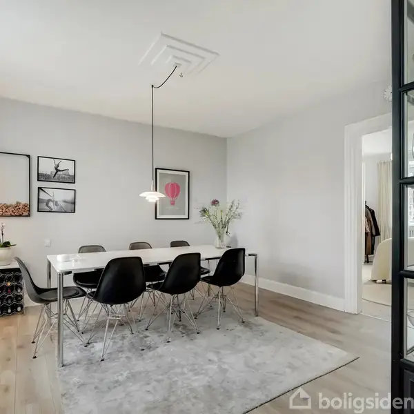 Et spisebord med seks sorte stole står på et lyst tæppe. To lamper hænger over bordet. Rummet har en åbning til en anden del af huset.