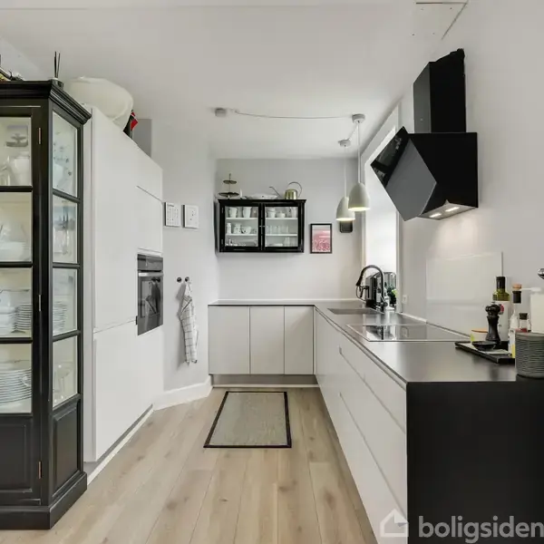 Et moderne køkken med hvide skabe og en sort glasvitrine til venstre indeholder porcelæn. En vask og kogeplader er på en mørk bordplade. Rummet er lyst og minimalistisk med trælaminatgulv.