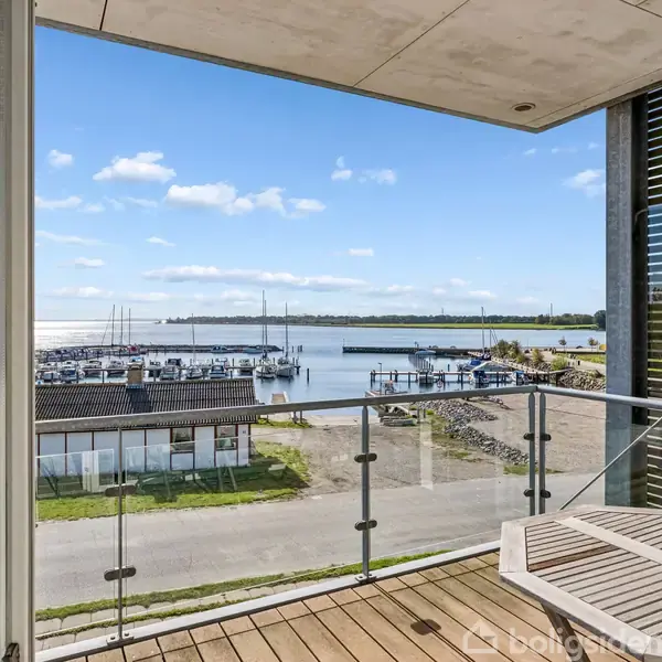 Balkon med glasrækværk, stående over en marina med både for anker i baggrunden på en solrig dag. Udsigt over vejen og vandet under en klar blå himmel.