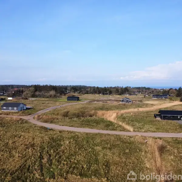 En række sommerhuse står spredt i et grønt, åbent landskab med en snoet asfaltvej. Husene har mørke tage og er omgivet af marker og træer under en klar blå himmel.