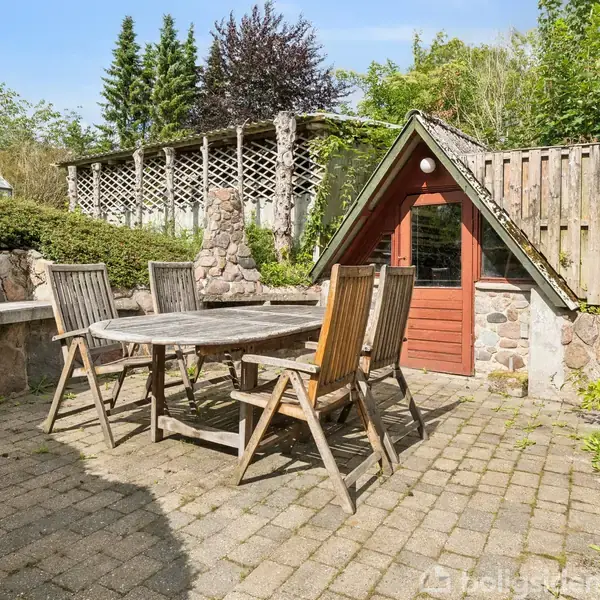 Et træbord omgivet af seks stole står på en brolagt terrasse. En lille bygning med en rød dør og stenvægge ligger tæt ved. Grønne træer omkranser området.