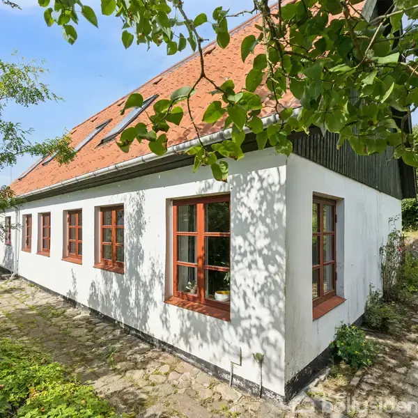 Et hvidt hus med rødt tegltag og røde vinduesrammer står på en stenbelagt sti. Det er omgivet af grønne træer og buske.