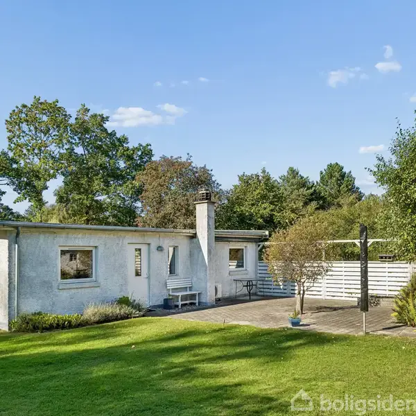 En hvid bungalow omgivet af en græsplæne og træer. Terrassen har havemøbler, og området er indhegnet med et lavt hegn. Himmel er klar og solrig.