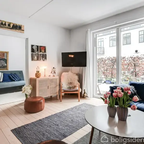 Stuen har en blå sofa, et rundt bord med tulipaner og en fladskærm på væggen. Store vinduer med udsigt til gårdhave omkranser rummet, der har lyst trægulv.