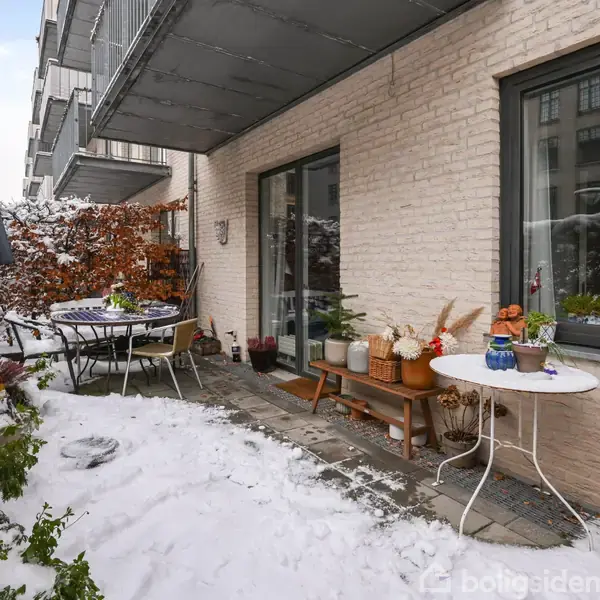 Terrasse dækket af sne med havemøbler og planter arrangeret omkring. En bygning med beige murstensvægge og vinduer danner baggrunden. Situationen udstråler en kold, vinterlig atmosfære.