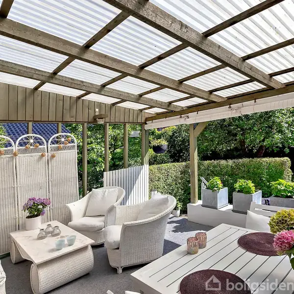 Terrasse med hvide kurvemøbler står under overdækning, omgivet af grønne planter. En sofagruppe med puder og et bord med dekorative stearinlys skaber en hyggelig atmosfære.