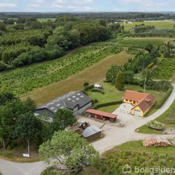 Gården ligger i et landligt område med et hovedhus i gul mursten, et stort gråt udhus og marker med skov i baggrunden. Veje omgiver ejendommen til venstre.
