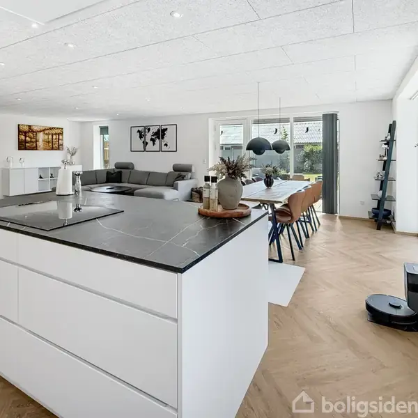 Køkkenø med sort bordplade står centralt, flankeret af grå sofaer og træspisebord med stole. Store vinduer giver naturligt lys. Moderne indretning med planter og dekorationer i et åbent rum.
