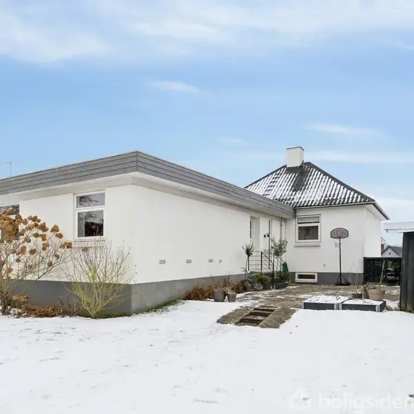 En hvid villa står stille omgivet af sneklædt have med et par buske og træer. En sti fører til en terrasse ved siden af huset.