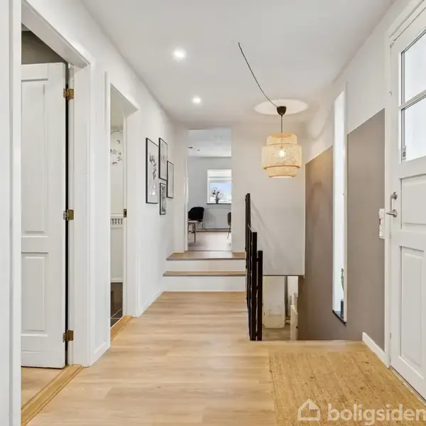 Dør åbner til lys gang med trægulv og væghængt lampe. Trapper fører ned til rum med skrivebord. Neutrale vægge, hvid dør med rude.