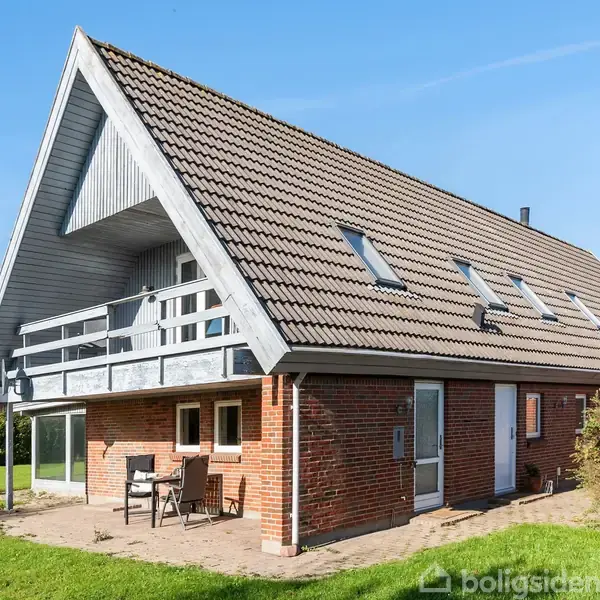 Et rødt murstenshus med en stor skråtag og loftskviste, står i en grøn have. Foran huset er en terrasse med borde og stole, omgivet af græsplæne og buske.