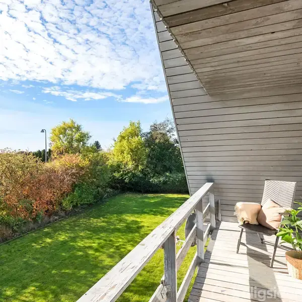 En træterrasse med en stol og plante står ovenpå en velplejet græsplæne, omgivet af buske og træer under en skyfyldt himmel.