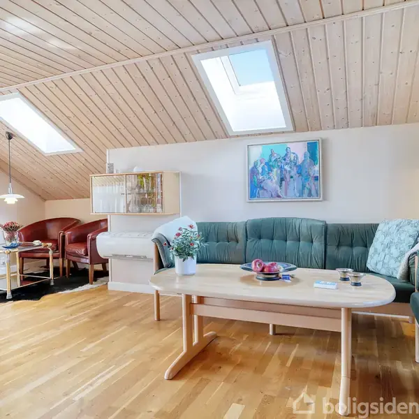 En stue med grøn sofa og sofabord i træ. To ovenlysvinduer oplyser rummet. I baggrunden er der en bar med flasker og nogle røde lænestole. En moderne maleri hænger på væggen.