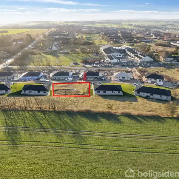 Grunde markeret med rødt ligger mellem moderne huse i et grønt landskab. Boligområder og marker strækker sig ud i det fjerne under en klar, blå himmel.