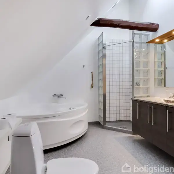 Badeværelse med skrå loft. Funktioner inkluderer et hjørnebadekar, brusekabine med glasvæg og en vask med spejl. Mørke skabe med belysning. Træbjælke synlig i loftet.
