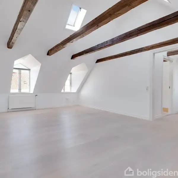 Et rummeligt, tomt loftsværelse med synlige træbjælker. Sollys strømmer ind gennem vinduerne og en ovenlysåbning. Til venstre ses radiator, og til højre er der adgang til køkkenet.