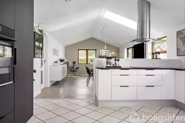Køkken med hvide skabe og en stor emhætte over kogepladen. Stuen ses i baggrunden med en skrånende loftsstruktur og vinduer, der slipper naturligt lys ind.