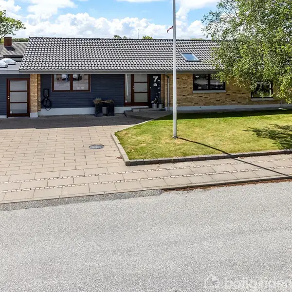 Et fritliggende hus med en grå facade og skråt tag, placeret ved en vej. Foran er en flisebelagt indkørsel og en velplejet græsplæne med et flagstang.