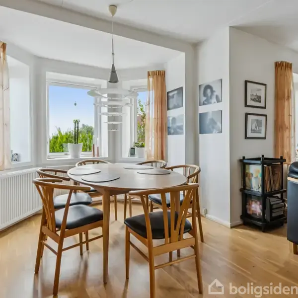 Et rundt spisebord med fire stole står i en lys spisestue. Store vinduer med lyse gardiner lader dagslys trænge ind. Væggene har forskellige sort-hvide billeder.