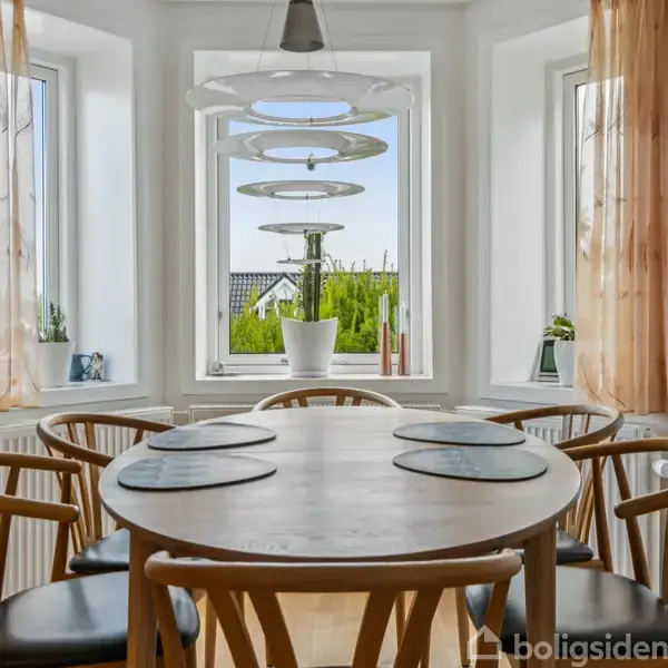 Rundt spisebord med seks stole, dækket med bordskånere, placeret i en spisestue med store vinduer. Udsigt til have og moderne lysarmatur hænger over bordet.