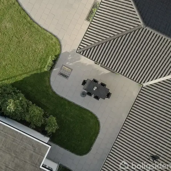Udendørs bord og stole står på en flisebelagt terrasse mellem to bygninger. Græsklædt område og buske omgiver terrassen i et moderne boligområde.