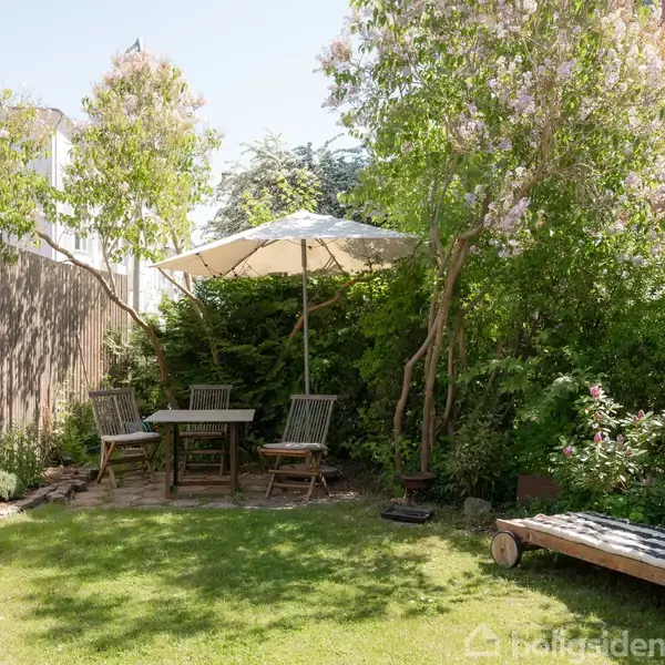 Havemøbler står under en parasol med et bord omgivet af stole. I baggrunden er der tætte buske og blomster, mens en liggestol står til højre i den solbeskinnede have.