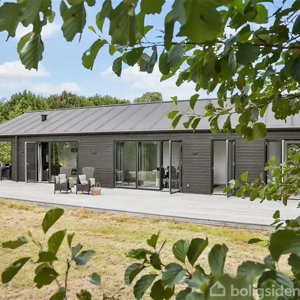 Et moderne, sort træhus med store glasdøre står stille på en terrasse i et grønt, træomkranset område. Der er havemøbler foran huset.