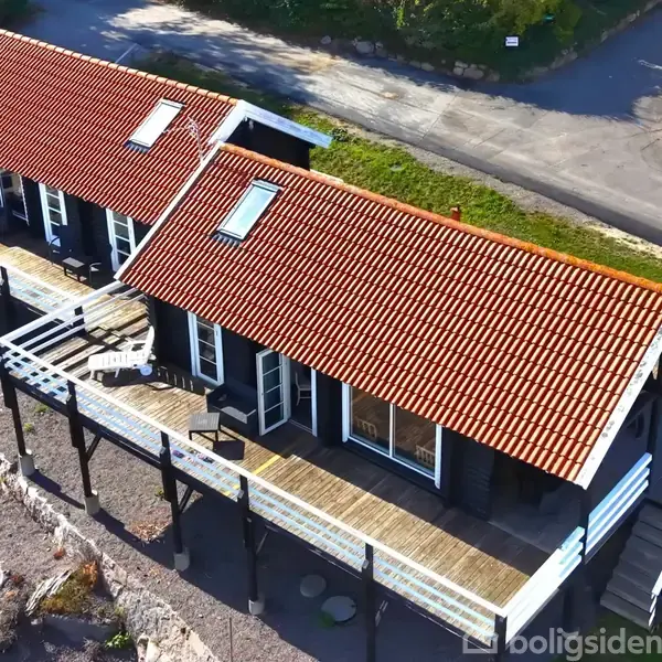 To sammenhængende sommerhuse med røde tegltage står på en hævet terrasse. De er omgivet af en grøn have og en asfaltvej i baggrunden.