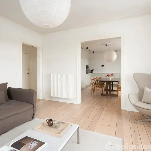 Grå sofa og lænestol står i et lyst stueområde med trægulv. Et sofabord med en bakke foran. Spisestue ses i baggrunden gennem en bred åbning.