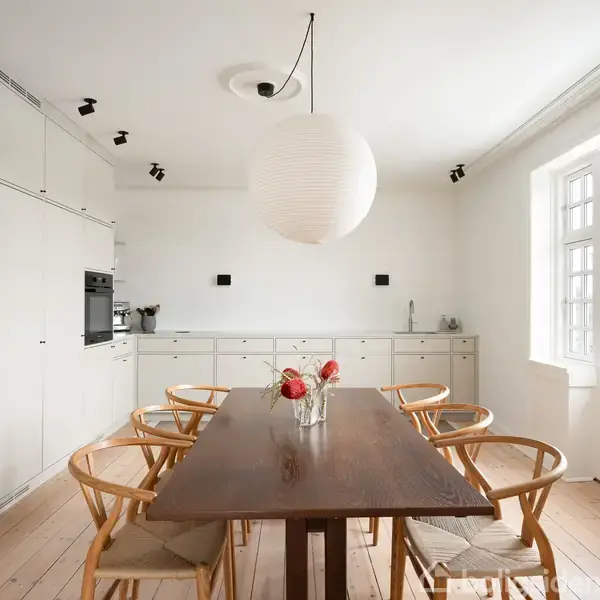 Et lakeret, mørkt træbord er omgivet af seks lyse træstole med lyse puder i et hvidt køkken. En stor, rund lampe hænger over bordet. Knappe lyspunkter ses i loftet.