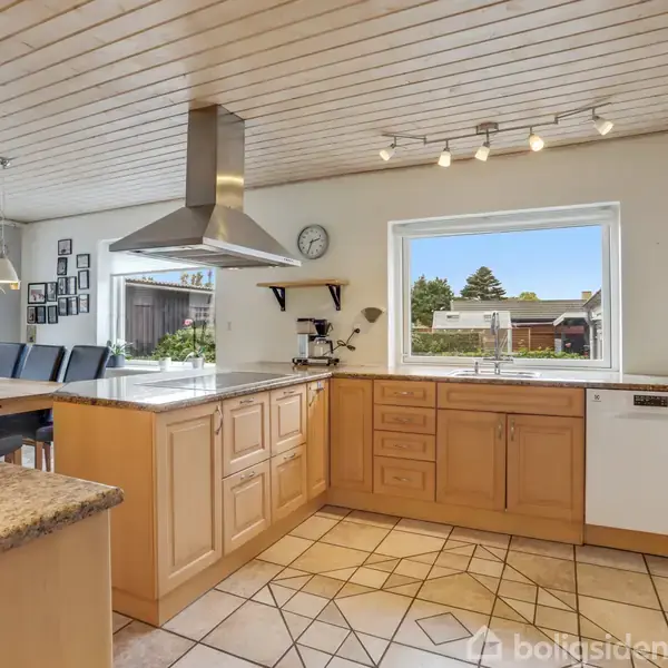 Et rummeligt køkken med træskabe og granitbordplader. En emhætte hænger over kogeøen. Indbygget ovn og opvaskemaskine ses. Stort vindue giver udsigt til en terrasse og have.