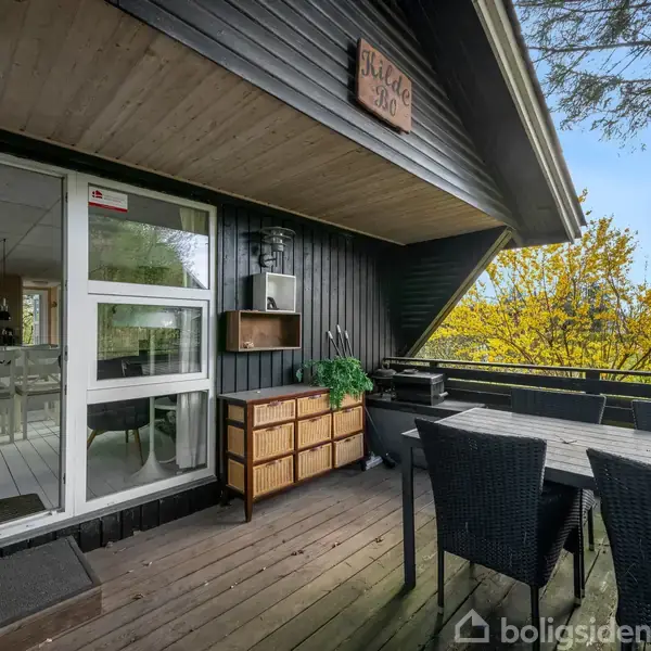 Træterrasse med sorte havemøbler og dekorativt skilt med teksten "HILDE SKOV". Terrasse ligger ved et hus, omgivet af grønne trætoppe, med udsigt til et lyst opholdsrum gennem glasdøre.