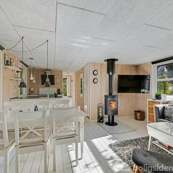 En stue med en brændeovn i hjørnet. En stor, hvid spisebord med stole står i forgrunden. Sofa og TV på væggen, omgivet af træpaneler og store vinduer.
