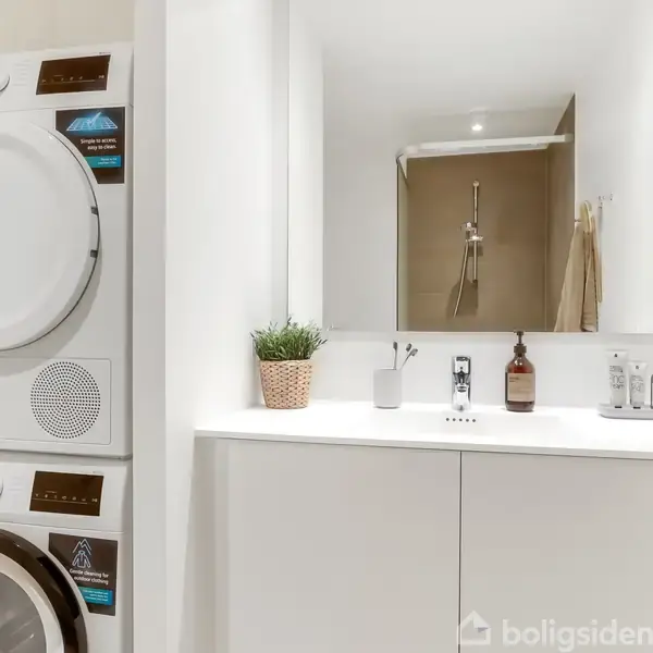 Vaskemaskine og tørretumbler står stablet til venstre i et moderne badeværelse. Håndvask med spejl og toiletartikler ses foran. I baggrunden er der en bruseniche.