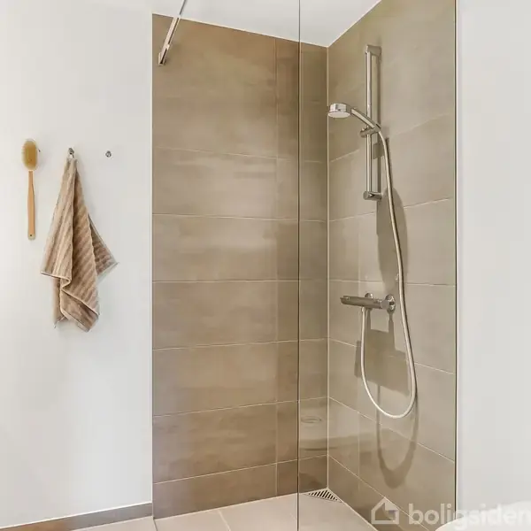 Bruseren er monteret på en væg med beige fliser og glasvæg. På modsat væg hænger et håndklæde og en børste. Rummet er lyst og minimalistisk.