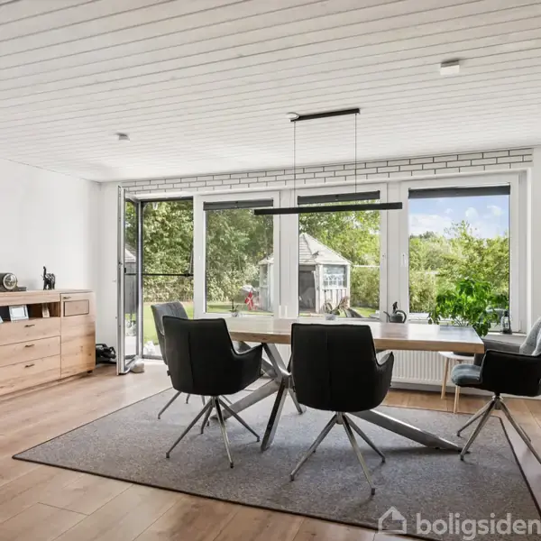 Fem moderne stole står omkring et træbord i en spisestue med stort vindue, der viser en grøn have. Skænk og loftlampe supplerer det lyse, luftige rum.