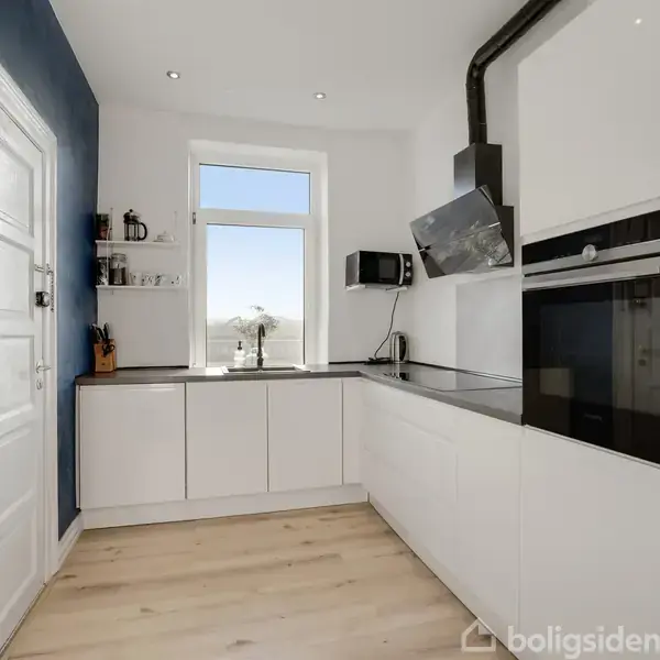 Et køkken med hvide skabe danner et hjørnearrangement. En sort emhætte er monteret over en kogeplade. Vinduet lader lys ind, og væggen er blå. En plante hænger på radiatoren.