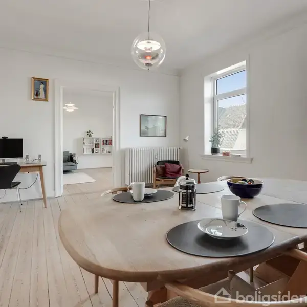 Et lyst spisebord med dækkede tallerkener og en kaffekande står i centrum. Baggrunden har en computerplads, reoler og store vinduer, der oversvømmer rummet med naturligt lys.