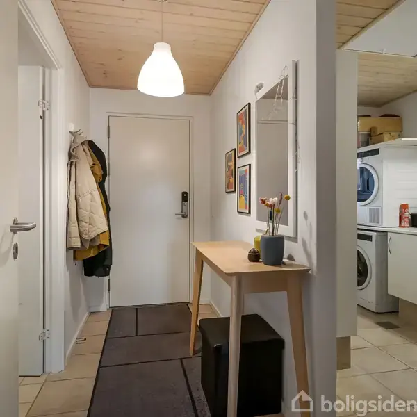 Gang med dør til venstre og frakkeophæng. Til højre står et træbord med blomster og spejl over. Bagved vaskerum med vaskemaskine, tørretumbler og skabe. Ingen synlig tekst bortset fra "boligsiden" vandmærke.