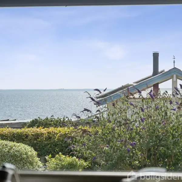 Udsigt fra et vindue viser en have med blomster og buske i forgrunden, med et lille hus bagved og havet i baggrunden under en klar blå himmel.