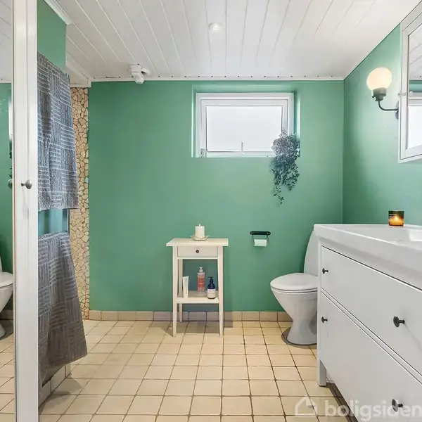 Badeværelse med grønne vægge og lyse fliser. Vask med spejl til højre, toilet til venstre, og lille hvidt bord i midten. Et vindue lader lys komme ind.