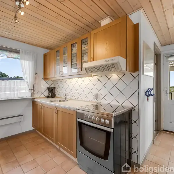 Køkken med træskabslåger og komfur, flisebelagt væg og lys flisegulv; vindue mod haven og dør, som åbner til terrasse. Hvidt loft med spotlys.