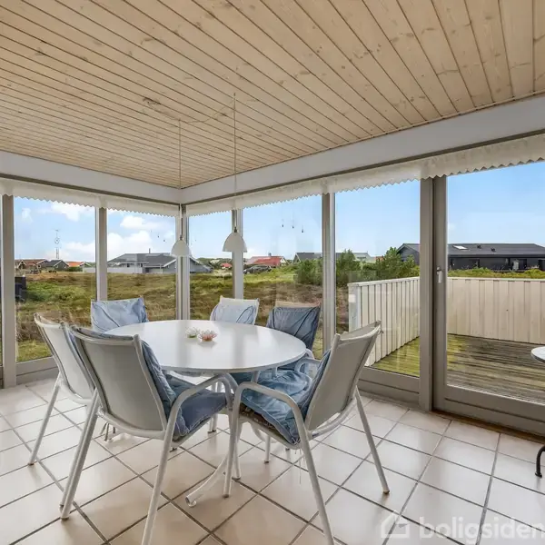 Rundt bord med fire stole står i et lyst rum med store vinduer, som giver udsigt til en terrasse og grønne omgivelser. Lyse fliser på gulvet og træloft kompletterer rummet.